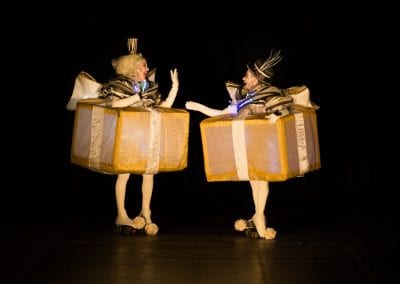 Two people in elaborate costumes shaped like large gold and white gift boxes, adorned with lights, facing each other in a dark setting.