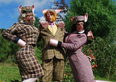 Three people wearing pig costumes with tweed suits pose outdoors in a garden with trees and grass under a blue sky.