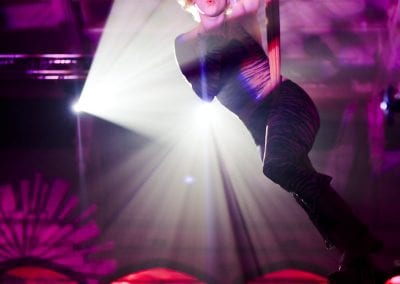 Performer with curly hair and dark outfit, suspended from aerial silks, in a spotlight. Background features red lighting and decorative elements.