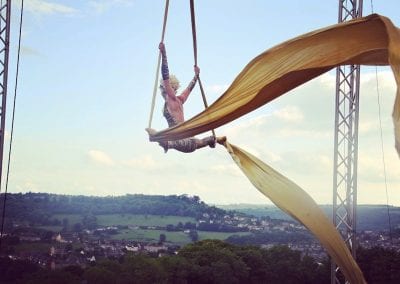 A person performs aerial silk acrobatics, hanging between two poles with a scenic landscape in the background.