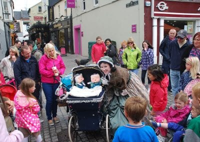 A street performer dressed in vintage clothing entertains a crowd by a double pram with two puppet babies. People of all ages gather and watch on a busy pedestrian street.
