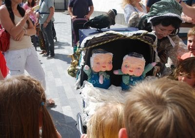 Children gathered around a puppet show featuring two puppet characters in a decorated stroller, watched by a nearby adult.