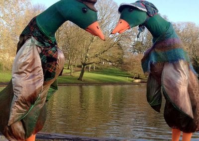 Two people in elaborate duck costumes with colorful scarves and hats stand by a pond, facing each other.