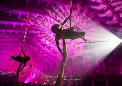 Two aerial performers in dramatic lighting, suspended on silks in a large venue with purple lighting and decorative string lights.