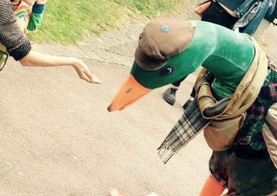 Person in a duck costume wearing a hat and scarf, bending forward on a path, while a person offers a hand nearby.