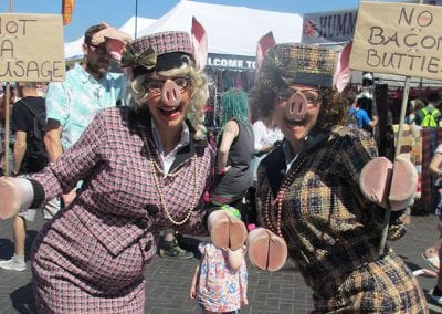 Two people in pig costumes hold signs reading Not a Sausage and No Bacon Butties! at an outdoor event, with a crowd in the background.