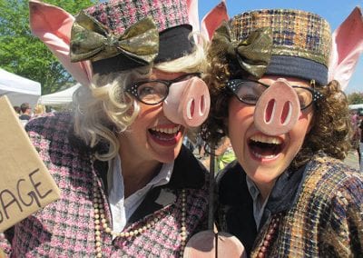Two people dressed in pig costumes, wearing glasses and plaid outfits, smile at an outdoor event.