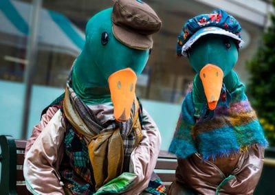 Two figures dressed as ducks sit on a bench, wearing colorful scarves, clothing, and hats, with building and trees in the background.