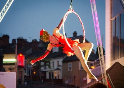 A performer in red costume performs on an aerial hoop at an outdoor event, with buildings and a darkening sky in the background.