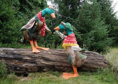 Two large duck costumes with colorful scarves and hats on a fallen log in a wooded area.