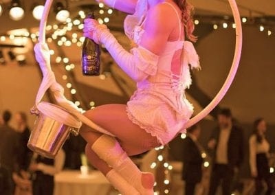 Performer in a white costume sits on a hoop, holding a bottle. She is elevated at an indoor event with lights and people in the background.