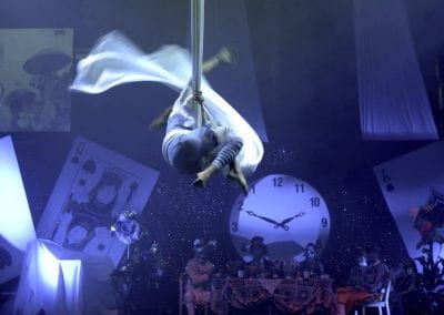 Aerialist performs on silk ropes above a stage set with oversized playing cards and a clock, themed as Alice in Wonderland.