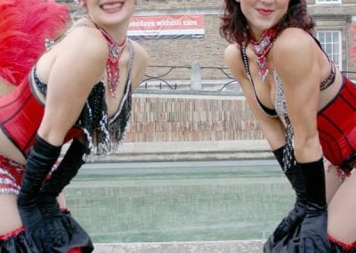 Two performers in ornate red and black costumes with feathered headpieces pose and smile in front of a brick building.
