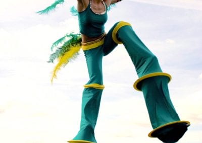 Person in a green costume with yellow and green feathers jumps energetically on stilts against a cityscape backdrop.