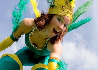 Performer in a green and yellow feathered costume, smiling and reaching forward, against a blue sky backdrop.