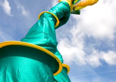 Person on stilts wearing vibrant green and yellow costume with tall, feathered headdress set against a blue sky.