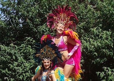 Two women in colorful carnival costumes with feathered headdresses stand outdoors. One is on stilts, wearing a long skirt with ruffles. Green foliage is in the background.