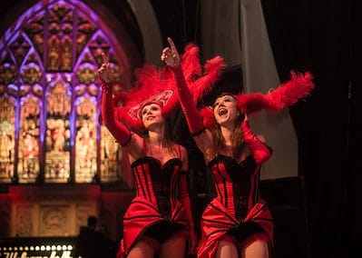 Two performers in red corsets and feathered hats gesture energetically on stage, seamlessly blending their craft with Circomedia flair against a stained glass backdrop.