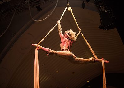 Aerial performer in a gold outfit executes a split pose while suspended on beige silks in an indoor venue.
