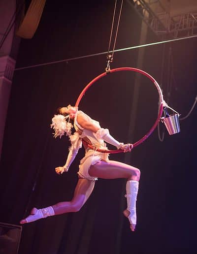 A performer in white costume does an aerial hoop act, with one leg extended, under stage lighting.