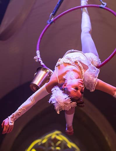 Aerial performer in white costume hangs upside down on a hoop, arms extended.