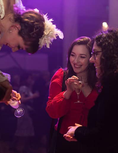 Person in costume pours champagne into glasses held by two women at an event with purple lighting.