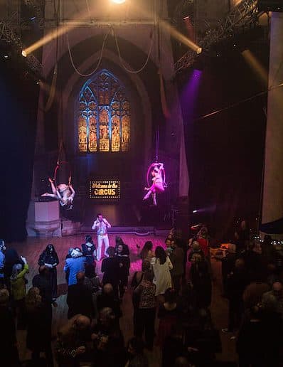 Performers on aerial hoops entertain a crowd in a dimly lit venue with stained glass windows and spotlight effects.