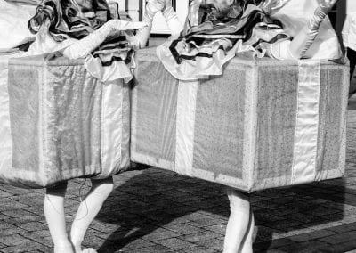 Two people dressed as gift boxes on roller skates, smiling and holding hands in an outdoor setting.