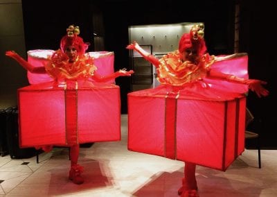 Two people in illuminated red box costumes with gold ribbons and matching headpieces, standing in a hallway.