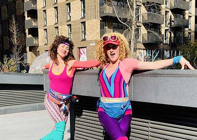 Two people in colorful outfits roller skating outdoors by a low wall on a sunny day, with buildings in the background.