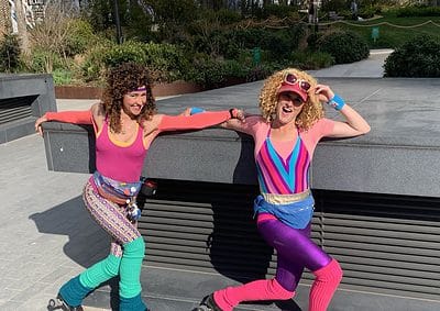 Two people in colorful retro outfits and roller skates pose playfully outdoors on a sunny day.