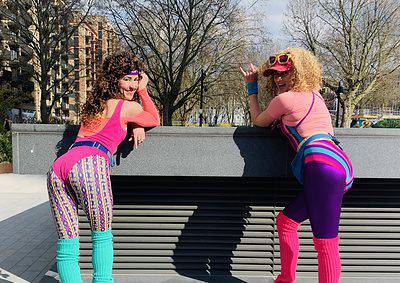 Two people in bright, retro clothing and roller skates pose playfully against a low wall outdoors on a sunny day.