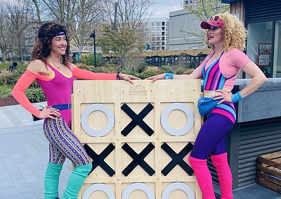 Two people in colorful retro workout outfits and roller skates stand beside a large wooden tic-tac-toe board in an outdoor urban area.