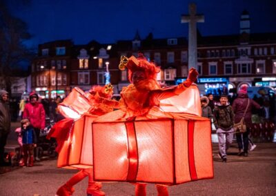 Performers from Cirqdelight, dressed as glowing gift boxes, enchant the crowd under the starlit sky in the bustling town square.