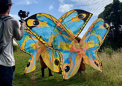 Person films another holding large, colorful butterfly-shaped kites under a cloudy sky with trees and grassy path.