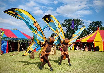 Two performers in brown costumes with large butterfly wings dance on grass near colorful tents under a blue sky.