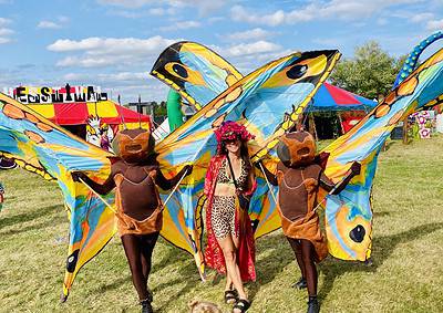 Three people in insect-themed costumes pose at an outdoor festival with colorful tents and a clear sky in the background.